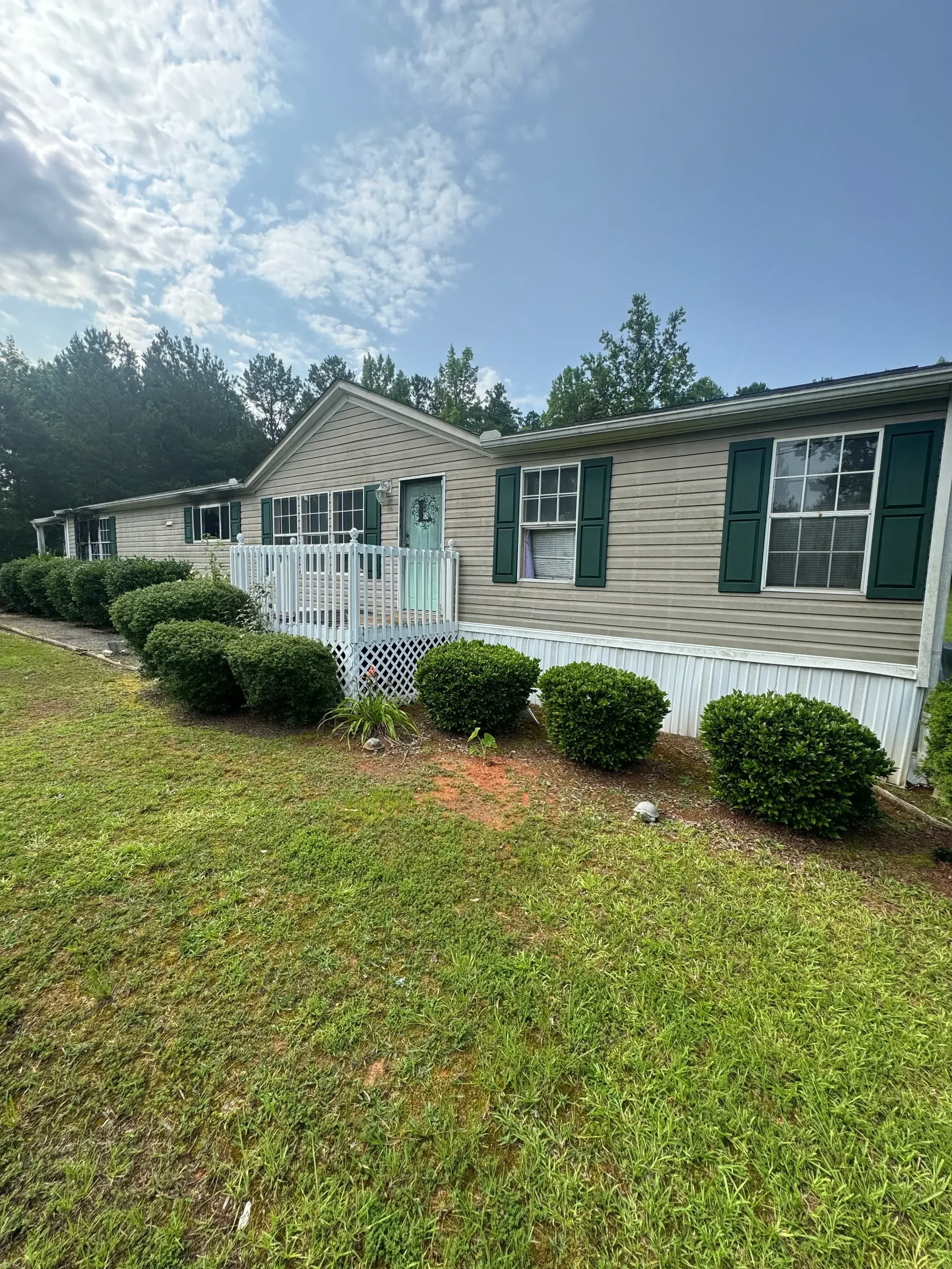 Cleared residential lot with manicured lawn, boxwood shrubs, and metal-sided house with dark green trim