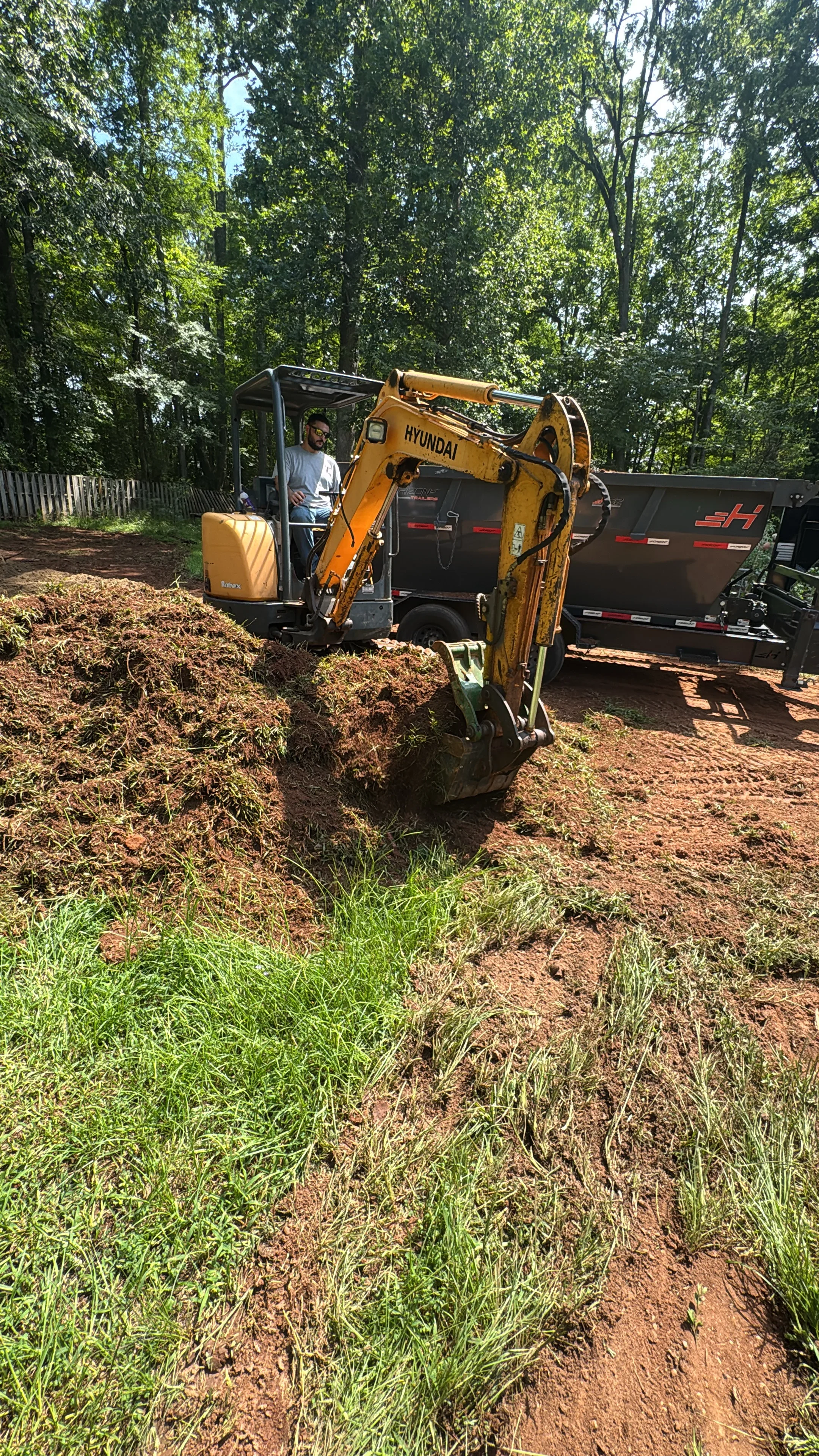 Mini excavator loading brush and debris into dump trailer on cleared residential lot